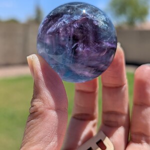 May include: A clear, spherical crystal ball with blue and purple hues is held in a hand. The crystal ball is the focal point, with a wooden letter "B" in the foreground. The background is blurred, with green grass and a blue sky.