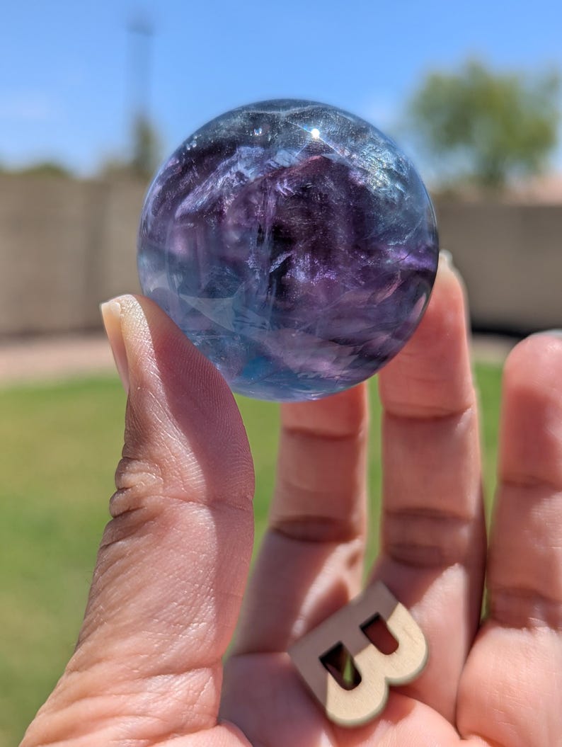 May include: A clear, spherical crystal ball with blue and purple hues is held in a hand. The crystal ball is the focal point, with a wooden letter "B" in the foreground. The background is blurred, with green grass and a blue sky.