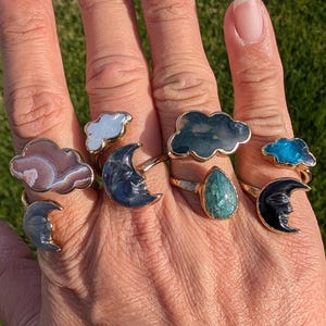 May include: A selection of gold-coloured rings with cloud and crescent moon motifs. The rings incorporate gemstones in shades of brown, white, blue, and green. The rings are displayed on a hand against a background of green grass.