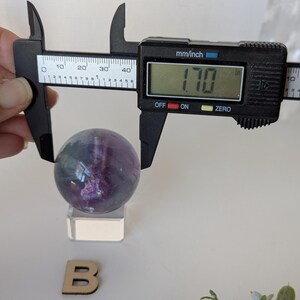May include: A purple and green fluorite sphere, approximately 1.7 inches in diameter, is measured with a digital caliper. The sphere sits on a clear acrylic stand. A wooden letter "B" is also present.