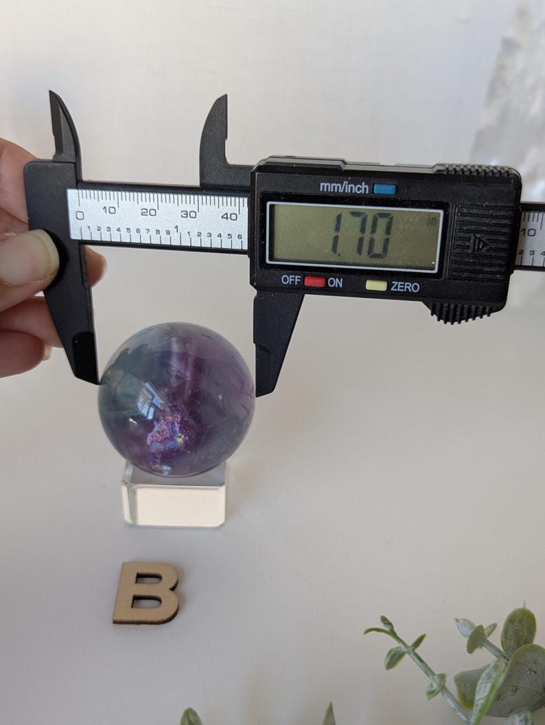 May include: A purple and green fluorite sphere, approximately 1.7 inches in diameter, is measured with a digital caliper. The sphere sits on a clear acrylic stand. A wooden letter "B" is also present.