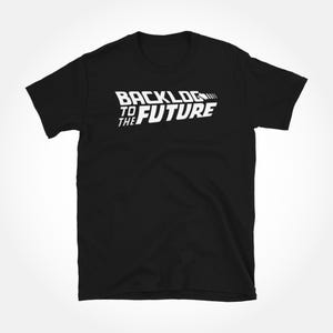 Puede incluir: Camiseta negra con el texto blanco "BACKLOG TO THE FUTURE" en una fuente estilizada. El diseño es un juego de palabras con el título de la película "Regreso al Futuro". La camiseta es de cuello redondo y parece estar hecha de algodón o un tejido similar.