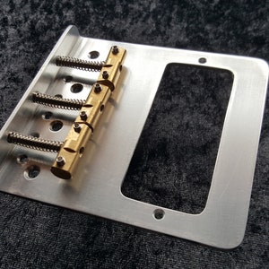 May include: A silver guitar bridge with four brass saddles and a rectangular cutout for a pickup. The bridge is designed for a standard 4-string electric guitar.