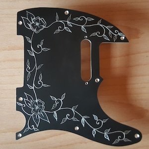 May include: Black guitar pickguard with a white floral design. The design features vines, leaves, and flowers.