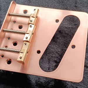 May include: A copper-coloured guitar bridge with brass saddles and springs. The bridge is rectangular with a curved cutout and multiple holes for mounting and string adjustment. The brass saddles are vertically aligned.