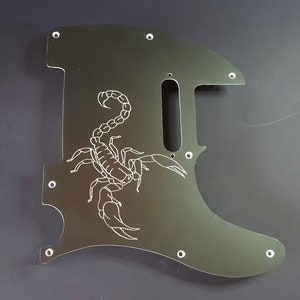 May include: A dark green guitar pickguard with a white scorpion design. The pickguard has a unique shape with several screw holes and a cutout for the pickups. The scorpion is intricately detailed.