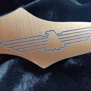 Puede incluir: Una placa de metal de color dorado con un acabado cepillado y una silueta negra de un águila estilizada con las alas extendidas.