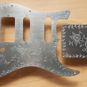 May include: Two silver metal guitar pickguards with an etched floral design. The larger pickguard is shaped like a guitar body and the smaller pickguard is rectangular.