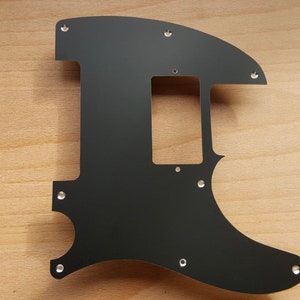 May include: A black guitar pickguard with a unique shape, featuring a rectangular cutout and several screw holes. The pickguard is made of a solid, matte black material and is designed to fit a specific guitar model.