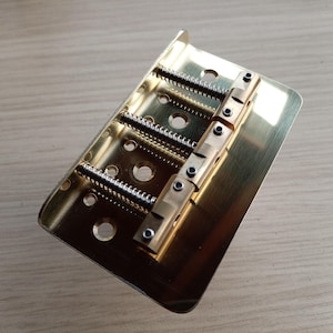 May include: A gold-colored guitar bridge with adjustable saddles and string height screws. The bridge has a rectangular base with multiple holes and is designed for a six-string instrument. The metal components are shiny and reflect light.