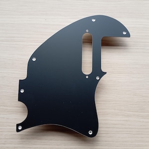 May include: A black guitar pickguard with a unique shape, featuring several screw holes and a rectangular cutout. The pickguard is made of a smooth, solid material and is designed to protect the guitar's body.