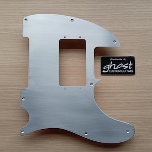 May include: A brushed silver guitar pickguard with a rectangular cutout and several screw holes. A black label with the text "ghost CUSTOM GUITARS" is also present. The pickguard is designed for a guitar.