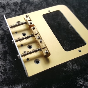 Puede incluir: Un puente de guitarra de color dorado con tres selletas ajustables y un recorte rectangular para una pastilla. El puente está sobre una superficie negra.