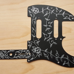 May include: Black guitar pickguard with a white floral design. The pickguard is made of metal and has a smooth, glossy finish.