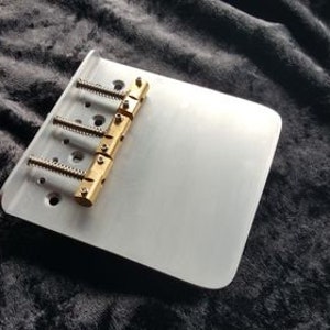 May include: A silver guitar bridge with four gold saddles. The bridge is designed for a four-string guitar.