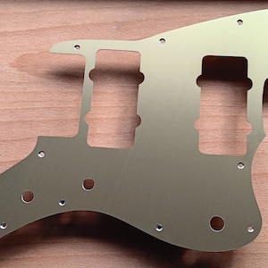 May include: A gold-colored guitar pickguard with various holes for pickups, knobs, and screws. The pickguard has a unique shape with a curved lower edge and a pointed upper section. It is made of a smooth, metallic material.