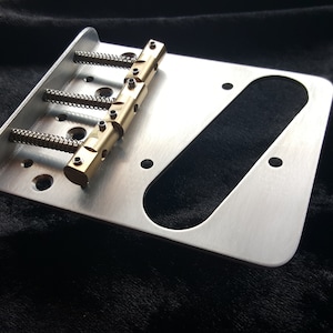 May include: A silver guitar bridge with four brass saddles and a cutout for a pickup. The bridge is designed for a six-string guitar.