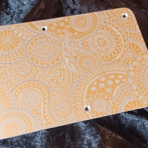 May include: A rectangular, gold-colored metal plate with a detailed, white paisley pattern. The plate has rounded corners and six evenly spaced holes. The design features intricate swirls and floral motifs.