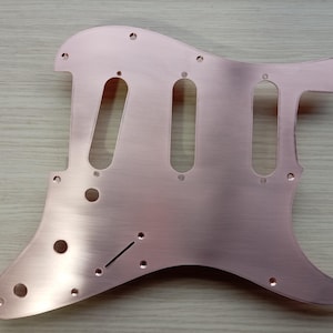 May include: A copper-colored guitar pickguard with a brushed metal finish. It has cutouts for pickups, knobs, and a tremolo arm. The pickguard is designed to fit a Stratocaster-style guitar.
