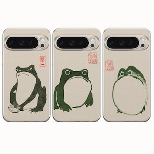 May include: Three beige smartphone cases featuring green frog illustrations. The first case shows a frog with a bowl, the second a seated frog, and the third a round frog. Each case includes red stamp-like text symbols in a distinct artistic style.