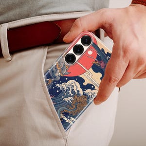 May include: A smartphone case featuring a graphic print of a large red sun, stylized blue ocean waves, and a dark bird silhouette. The case has three circular camera lens cutouts at the top and is partially inserted into a light-colored trouser pocket.