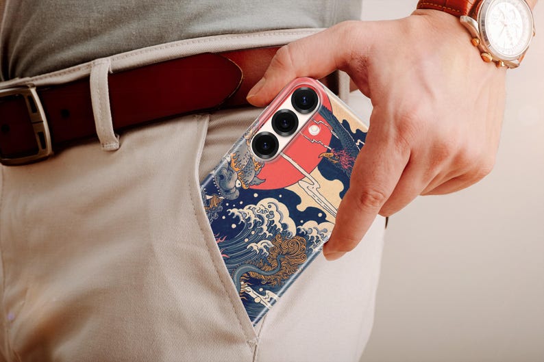 May include: A smartphone case featuring a graphic print of a large red sun, stylized blue ocean waves, and a dark bird silhouette. The case has three circular camera lens cutouts at the top and is partially inserted into a light-colored trouser pocket.