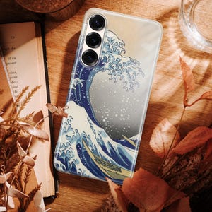 May include: A smartphone case featuring a graphic print of a large blue ocean wave with white foam crests, reminiscent of traditional Japanese woodblock art. The case has three circular camera lens cutouts positioned at the top left corner.