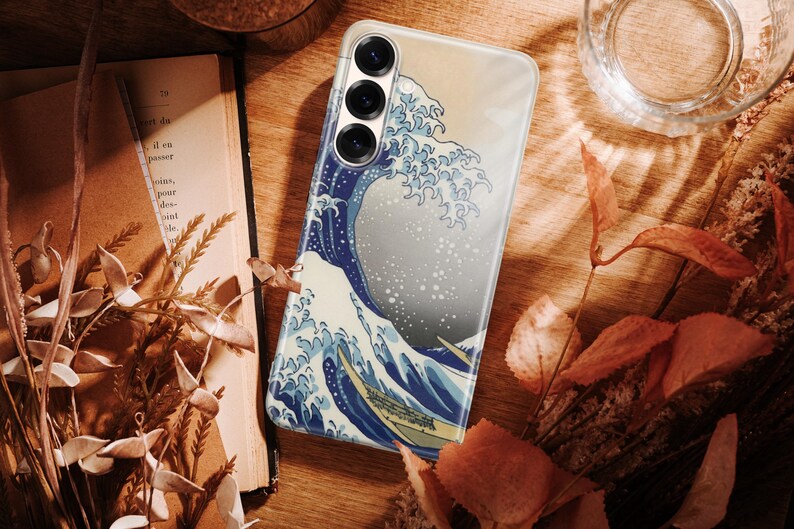 May include: A smartphone case featuring a graphic print of a large blue ocean wave with white foam crests, reminiscent of traditional Japanese woodblock art. The case has three circular camera lens cutouts positioned at the top left corner.