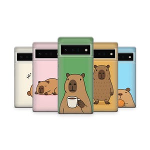 May include: A collection of five phone cases with capybara illustrations. Each case features a different color: cream, pink, green, yellow, and blue. The capybaras are depicted in various poses, including holding a coffee cup and sleeping.