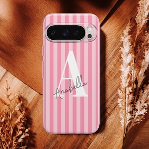 Puede incluir: Funda de teléfono a rayas rosas y blancas con el nombre "Anabella" en cursiva y una gran "A" blanca. La funda tiene un recorte para la cámara y está sobre una superficie marrón con plantas decorativas.