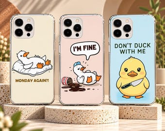 May include: Three clear smartphone cases featuring duck illustrations. The left case is cream with a duck on a pillow and the text MONDAY AGAIN?!. The middle case is pink with a duck, chocolate, and the text I'M FINE. The right case is light blue with a duck holding a knife and the text DON'T DUCK WITH ME.
