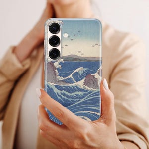 May include: A smartphone case featuring a printed reproduction of a Japanese woodblock print depicting turbulent blue ocean waves, rocky cliffs, and a distant shoreline under a pale sky, with three circular camera lenses visible at the top.