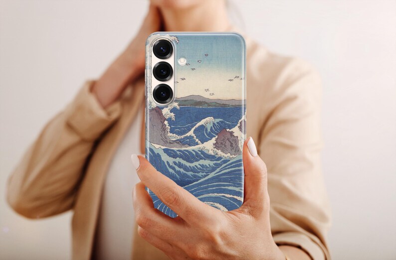 May include: A smartphone case featuring a printed reproduction of a Japanese woodblock print depicting turbulent blue ocean waves, rocky cliffs, and a distant shoreline under a pale sky, with three circular camera lenses visible at the top.