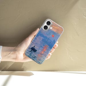 May include: A clear smartphone case featuring a printed reproduction of an impressionist painting depicting a harbor scene at sunrise with a small boat, blue water, and a bright orange sun reflecting on the surface, with two camera lens cutouts.