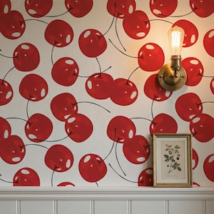 May include: A wall covered in a repeating pattern of red cherries on a cream background. A brass wall sconce with a lightbulb is mounted on the wall. A framed botanical print is positioned below the sconce.