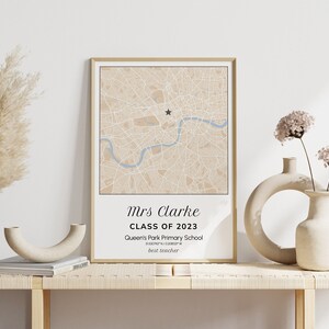 May include: A framed map print with a beige and blue color scheme, featuring a star marker and text that reads "Mrs Clarke, Class of 2023, Queen's Park Primary School, best teacher." The print is displayed on a light wooden table with decorative vases.