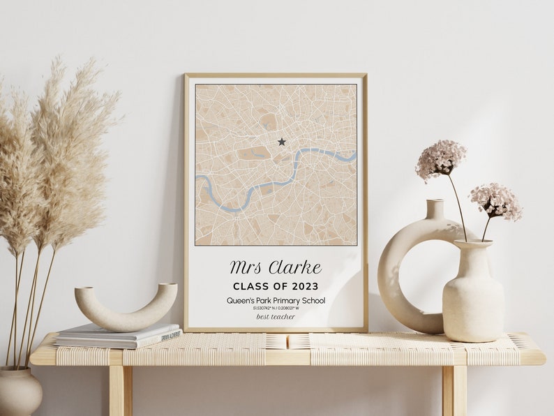 May include: A framed map print with a beige and blue color scheme, featuring a star marker and text that reads "Mrs Clarke, Class of 2023, Queen's Park Primary School, best teacher." The print is displayed on a light wooden table with decorative vases.