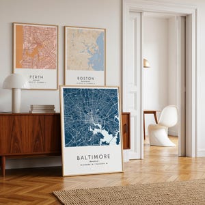 May include: Three framed city map prints. The maps are of Perth, Boston, and Baltimore. The Perth map is peach and brown, the Boston map is blue and tan, and the Baltimore map is navy blue and white. The Baltimore map is the largest and is leaning against a wooden cabinet.