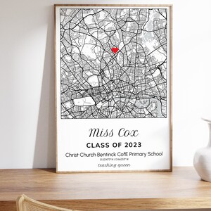 May include: A framed black and white map print with a red heart marking a location. The print includes the text "Miss Cox CLASS OF 2023" and school details. The print is displayed on a wooden surface.