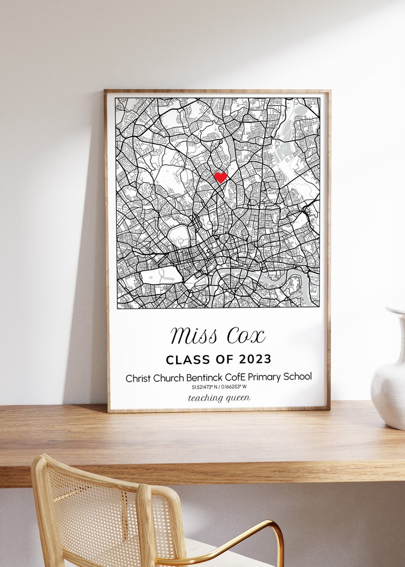 May include: A framed black and white map print with a red heart marking a location. The print includes the text "Miss Cox CLASS OF 2023" and school details. The print is displayed on a wooden surface.
