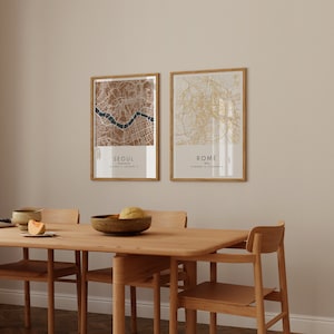 May include: Two framed maps of Seoul, South Korea and Rome, Italy, hanging on a beige wall above a wooden dining table with four chairs. The maps are in a minimalist style with brown and gold lines.