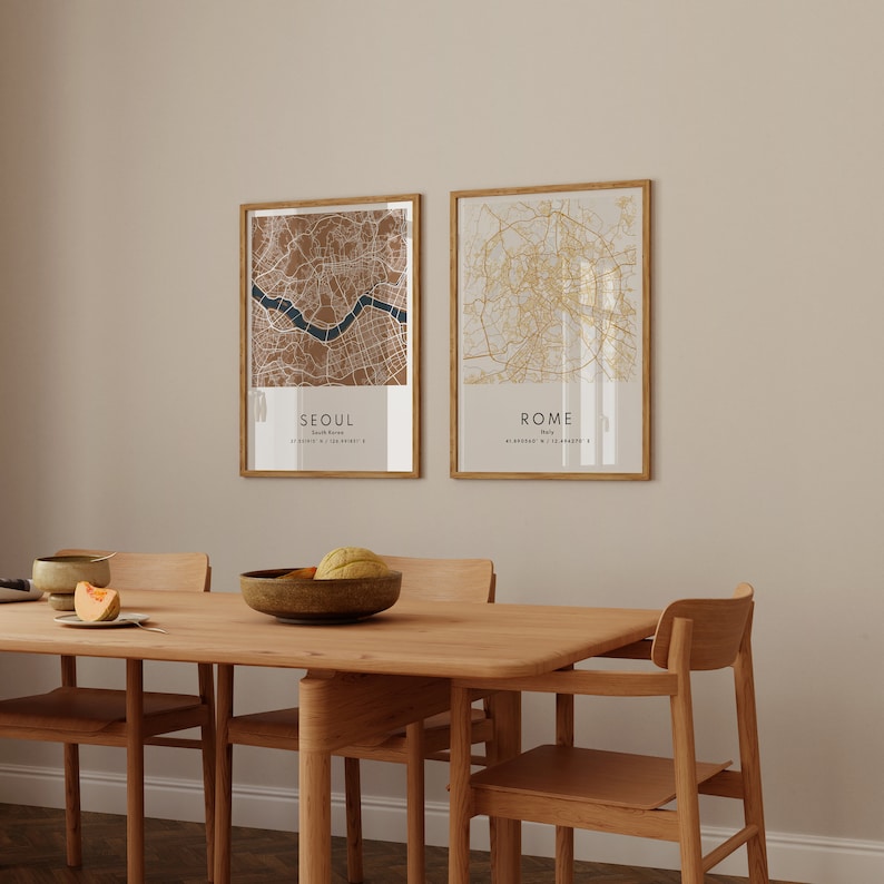 May include: Two framed maps of Seoul, South Korea and Rome, Italy, hanging on a beige wall above a wooden dining table with four chairs. The maps are in a minimalist style with brown and gold lines.