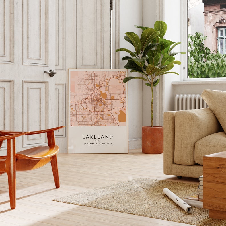 May include: A framed map print of Lakeland, Florida, with a pink background and brown lines showing the city streets. The map is hanging on a white wall in a living room setting with a brown chair, a potted plant, and a beige couch.