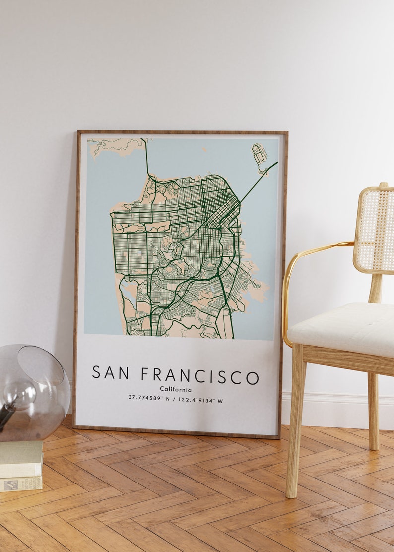 May include: A framed map of San Francisco, California, with a light blue background and green lines representing streets. The map is framed in a light brown wood frame and is hanging on a white wall. The text "SAN FRANCISCO" and "California" is printed below the map, along with the coordinates "37.774589&deg; N / 122.419134&deg; W".