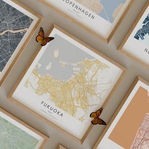 May include: A framed map print of Fukuoka, Japan, with a light gray background and gold lines representing streets. The map is surrounded by a light wood frame. Two orange butterflies are on the wall near the map.
