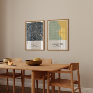 May include: Two framed city maps, one of Berlin, Germany and one of Milwaukee, Wisconsin, hanging on a beige wall above a wooden dining table with four chairs.