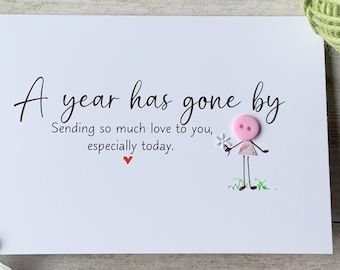 Op de afbeelding: Een witte kaart met de tekst "A year has gone by" en een liefdesboodschap. Een roze knoopfiguur houdt een bloem vast. Een klein rood hart staat onder de tekst. Een madeliefje en groen garen staan op de achtergrond.