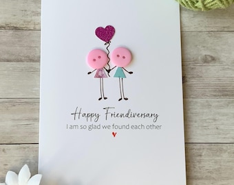 Op de afbeelding: Een witte kaart met de tekst "Happy Friendiversary I am so glad we found each other" en een roze hart. Twee roze knopen met getekende gezichten houden elkaar vast. Een roze hart met glitter is bevestigd aan een touwtje boven de knopen.