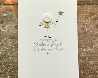 Op de afbeelding: Witte kerstkaart met een knoop engel illustratie. De engel heeft een goud glitter lichaam en houdt een sterrenstaf vast. De kaart leest "Sending you a Christmas Angel to look after you this year" met een rood hart.