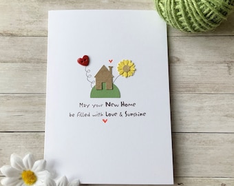 Op de afbeelding: Een witte kaart met een bruin huis, een groene heuvel, een gele zonnebloem en een rood hart. De kaart zegt "Moge je nieuwe huis gevuld zijn met liefde en zonneschijn".